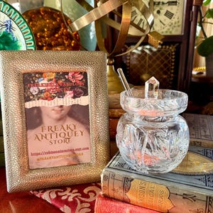 May include: A gold-toned textured picture frame with a vintage image and text for "Freaky Antiquey Store". A clear, cut-glass jar with a crystal lid and spoon sits on antique books. A brass armillary sphere and a clock are in the background.