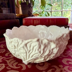 May include: A white ceramic bowl featuring a raised, textured leaf pattern across its exterior surface. The bowl has a scalloped, uneven rim and is supported by small, integrated feet at the base, creating a detailed, organic appearance.