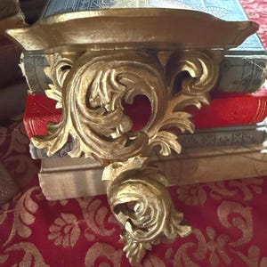 May include: A gold-colored, ornate wall shelf with a decorative, scrollwork design. The shelf is mounted above a stack of antique books with red, blue, and tan covers. The shelf has a small, curved lip.