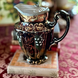 May include: An antique black and gold ceramic pitcher with a silver lid. The pitcher features floral and leaf designs with gold accents. The handle is black and curved. The pitcher is sitting on top of a stack of books.