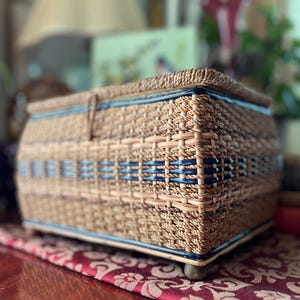 May include: Woven rectangular basket with a lid. The basket is made of light brown and tan woven material with blue accents. The basket sits on small wooden feet and is placed on a red patterned surface. The basket is likely used for storage.