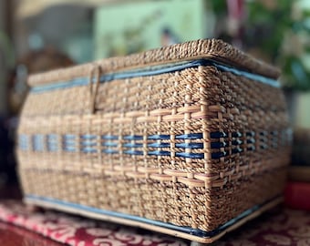 May include: Woven rectangular basket with a lid. The basket is made of light brown and tan woven material with blue accents. The basket sits on small wooden feet and is placed on a red patterned surface. The basket is likely used for storage.