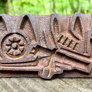 May include: A rectangular, weathered, cast iron object with a detailed relief design. The design includes a spoked wheel, architectural elements, and stylized floral or foliate motifs. The object has a rusty brown color and two small holes.