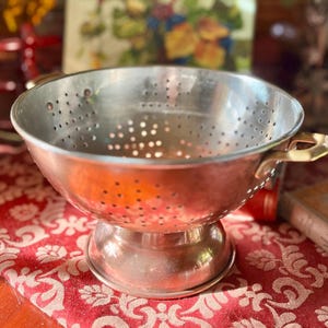 May include: A vintage metal colander with a perforated bowl and a pedestal base. The colander has two brass-colored handles and is sitting on a red patterned tablecloth. The bowl is silver-colored.