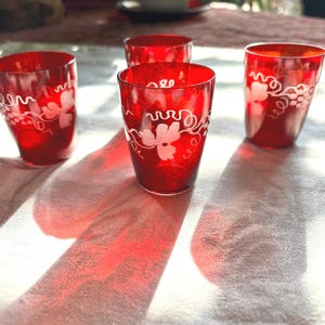 May include: Four red glass shot glasses with a white etched grapevine design. The glasses are on a white tablecloth.