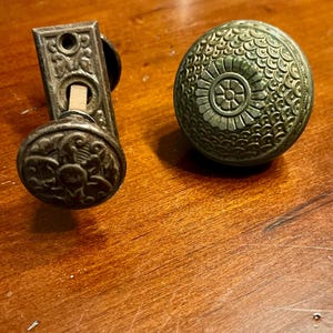 May include: Two antique door knobs. One is a rectangular plate with a round knob, the other is a round knob with a floral design. Both are made of a dark metal and sit on a wooden surface.