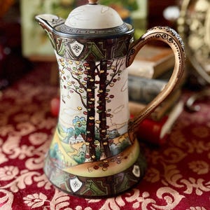 May include: A tall, cream-colored ceramic coffee pot with a lid and handle. The pot features a landscape scene with trees and a house, and is decorated with brown, green, and gold accents. The handle has a decorative pattern.