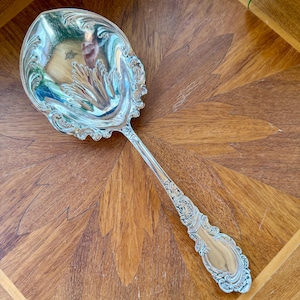May include: A silver serving spoon with an ornate, floral design. The spoon has a wide, oval bowl and a long handle with detailed scrollwork. The spoon is likely made of sterling silver and is a vintage piece.