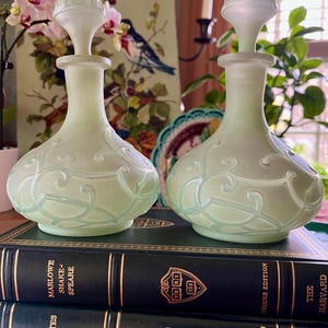 May include: Two vintage green glass perfume bottles with ornate designs and frosted glass stoppers. The bottles are sitting on a stack of three black hardcover books with gold lettering and a gold crest.