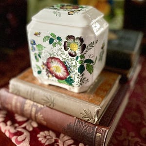 May include: A white ceramic jar with a lid, adorned with floral patterns in shades of red, purple, and green. The jar sits atop a stack of antique books with aged covers, resting on a patterned red fabric.