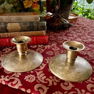 May include: Two antique brass candle holders with floral engravings. The candle holders have a wide, flat base and a small cup to hold the candle. The background includes a red tablecloth with a gold floral pattern, books, and a painting.