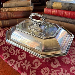 May include: A silver-colored, octagonal serving dish with a lid and handle. The handle is a geometric design. The dish is on a red patterned tablecloth, with antique books in the background.