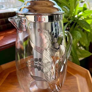 May include: A clear glass cocktail shaker with a silver metal lid and interior. The glass is decorated with a silver floral design. The shaker has a spout and a handle. The background includes a wooden tray and greenery.