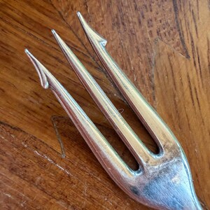 May include: Close-up of a silver fork with three tines. The fork is made of a shiny metal and has a simple, elegant design. The tines are pointed and slightly curved. The fork is resting on a wooden surface.