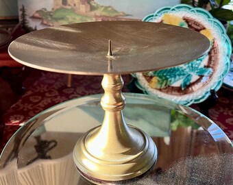 May include: A brass candle holder with a round base and a circular top. The candle holder has a classic design with a central spike for holding a candle. The brass has a warm, golden hue, and the piece is set on a reflective surface.