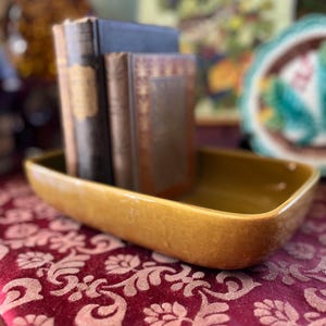 May include: A gold ceramic rectangular dish with rounded edges sits on a red patterned fabric. Three books are stacked inside the dish, including a dark blue book, a brown book, and a book with a gold design.