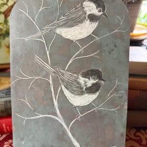 May include: A gray, arched plaque featuring two detailed bird illustrations perched on branches. The birds are depicted in black and white, with intricate feather and branch details. The plaque is supported by a black stand.