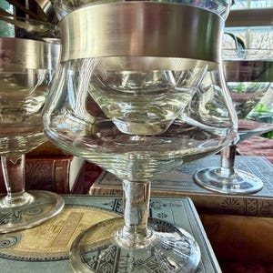 May include: Clear glass goblets with a silver band around the bowl. The goblets have a wide base and a stem. The goblets are sitting on top of antique books. The background includes a clock and a window.