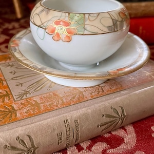 May include: A white porcelain teacup and saucer set. The cup and saucer have a gold rim and are decorated with orange floral designs and gold accents. The set is placed on top of a vintage book with gold lettering.
