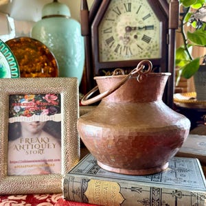 May include: A hammered copper vase with a handle sits atop antique books. The vase has a textured surface and a small handle. A framed print and a vintage clock are in the background. The scene evokes a vintage aesthetic.