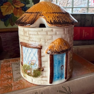 May include: A ceramic cottage-shaped jar with a thatched roof and a small chimney. The jar features a blue door, a window with a floral scene, and a textured brick-like exterior. The roof is brown and the jar is white.
