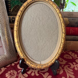 May include: An oval picture frame with a metallic gold finish and an ornate, embossed border pattern. The frame features a light gray, textured matting insert and is supported by a small, black, two-legged stand for upright display.
