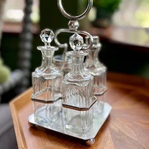 May include: A vintage silver-toned condiment set with four clear glass bottles. Each bottle has a faceted crystal stopper. The set sits on a silver tray with a handle and is displayed on a wooden tray.