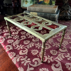 May include: A rectangular brass trivet with an openwork design featuring floral and scroll motifs. The trivet has four slender, rounded legs. The background includes a patterned red fabric and antique books.