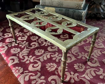 May include: A rectangular brass trivet with an openwork design featuring floral and scroll motifs. The trivet has four slender, rounded legs. The background includes a patterned red fabric and antique books.