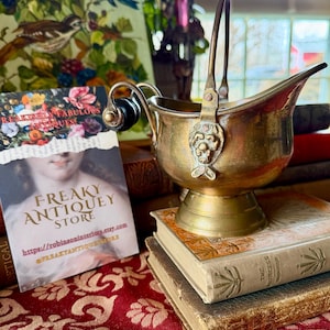 May include: A vintage brass coal scuttle with a handle and a decorative lion's head. The scuttle sits on a stack of antique books. A business card for "Freaky Antique Store" is visible.