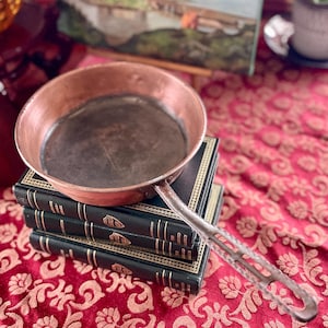 May include: A copper frying pan with a long, silver handle sits on top of a stack of four black hardcover books with gold lettering. The books are on a red tablecloth with a floral pattern.