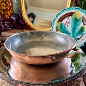 May include: A small copper pan with a single handle. The pan is sitting on a round, mirrored surface.