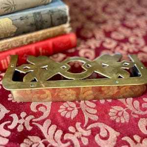 May include: A rectangular brass book holder with an ornate, openwork design. The holder has a decorative floral pattern and is resting on a red patterned surface. Several antique books are in the background.