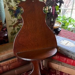 May include: A vintage wooden wall shelf with a dark brown finish. The shelf has a decorative, scalloped top and a small, curved shelf. The shelf is supported by a decorative base and is designed to be hung on a wall.