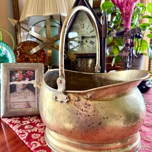 May include: A vintage brass coal scuttle with a curved handle. The scuttle has a rounded body with a flared rim and a decorative base. The surface shows signs of age and use. A framed picture and a clock are in the background.