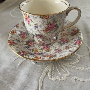 May include: A white teacup and saucer with a floral pattern in shades of pink, purple, yellow, and orange. The cup and saucer have a gold trim.