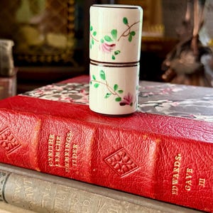 May include: A small cylindrical white porcelain object with a hand-painted floral vine pattern featuring green leaves and pink blossoms, accented by thin brown horizontal bands at the top, middle, and bottom edges of the two-part structure.