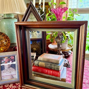 May include: A dark wooden framed mirror with a stack of antique books inside. The books have red, blue, and tan covers. A small framed picture with the text "FREAKY ANTIQUEY STORE" is on the left.