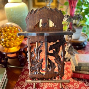 May include: A dark brown, intricately carved wooden display stand with a shelf. The stand features detailed carvings of birds and foliage. It has a small, rectangular shelf and is supported by four small legs. The stand is set on a red patterned fabric.