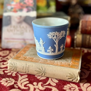 May include: A blue ceramic cup with white relief detailing, featuring classical figures and a tree design. The cup sits atop a vintage book with gold accents and is set against a blurred background with other books and a patterned tablecloth.