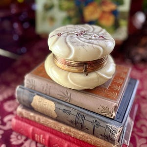 May include: A stack of antique books topped with a cream-colored, round trinket box. The box has a floral design on the lid and a gold-colored band. The books are various colors, including red, tan, and blue.