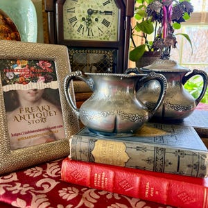 May include: An antique silver creamer and sugar bowl set with ornate detailing, resting on a stack of vintage books. A framed print with the text "FREAKY ANTIQUEY STORE" is visible. A wooden clock and a pink vase are in the background.