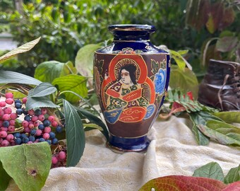 May include: A small blue and gold ceramic vase with a painted design of a woman in a red and gold robe. The vase has a round body and a narrow neck.