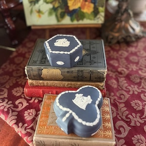 May include: Two blue decorative boxes with white relief designs, one hexagonal and one clover-shaped, rest on antique books. The boxes feature floral and cameo-style patterns. The background includes a painting and a patterned red tablecloth.
