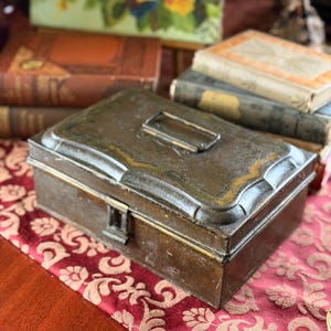 May include: A vintage metal box with a hinged lid and a small handle. The box is a dark, aged bronze color with gold trim and a clasp closure. It sits on a red patterned runner, with antique books in the background.
