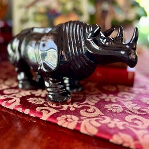 May include: A glossy black ceramic rhinoceros figurine with two horns and detailed skin texture. The rhino is positioned on a red patterned surface, suggesting a decorative or collectible item. The figurine has a reflective surface.
