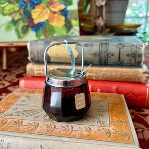 May include: A vintage, ruby-colored glass jar with a silver lid and handle, resting on antique books. The jar has a small spoon and a label that reads "Fine Quality". The background includes a painting and a potted plant.