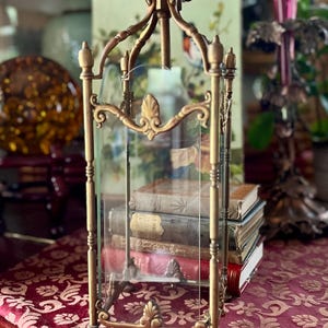 May include: An antique brass lantern with clear glass panels, showcasing a collection of vintage books inside. The lantern features ornate detailing and is suspended from a chain. The background includes a floral painting and other decorative items.