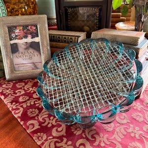 May include: A round, light blue glass flower frog featuring a scalloped edge and a silver-toned metal wire grid insert. The glass has a translucent quality with a smooth, reflective surface, designed to hold floral stems upright in a decorative arrangement.