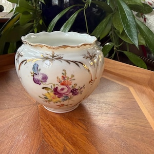 May include: A white porcelain bowl with a scalloped rim and gold trim. The bowl is decorated with colorful floral designs in shades of purple, pink, yellow, and blue. The bowl sits on a wooden tray.